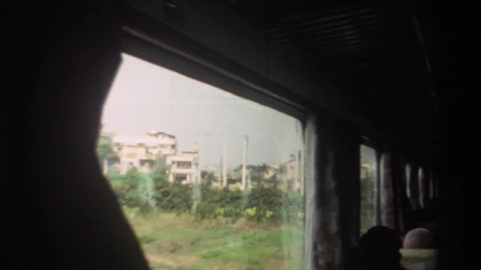 Inside a train in Taiwan, 1982, dark passenger silhouettes frame a window view of low buildings, poles, and greenery.