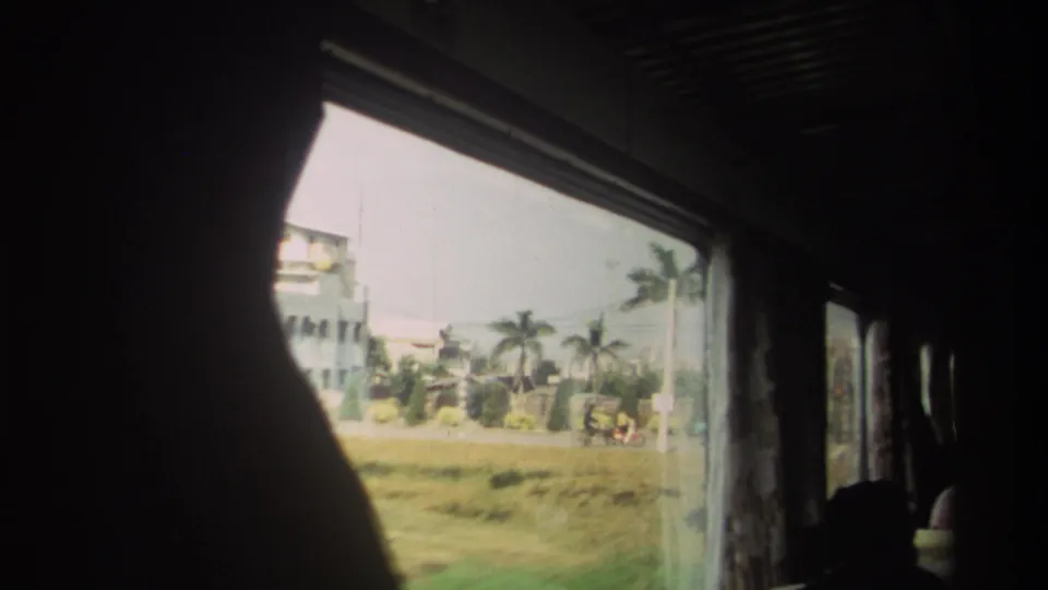 The window frames palms, grass, low buildings, and small roadside vehicles in Taiwan, with the bus interior remaining mostly dark.