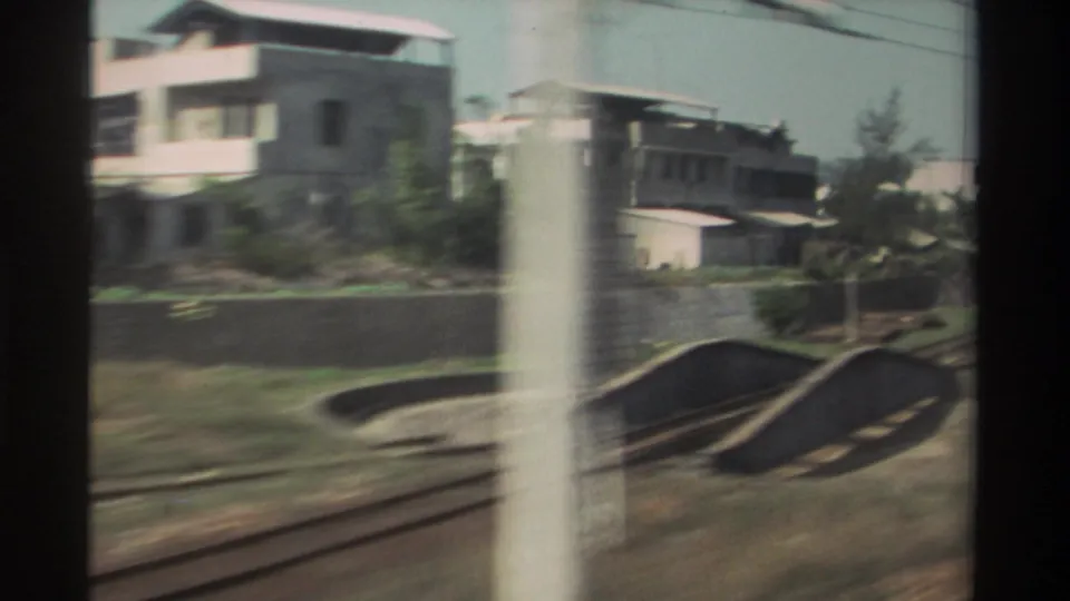 1982 Taiwan view from a moving train, showing railway tracks, a low wall, residential buildings, trees, and a blurred pole in front.