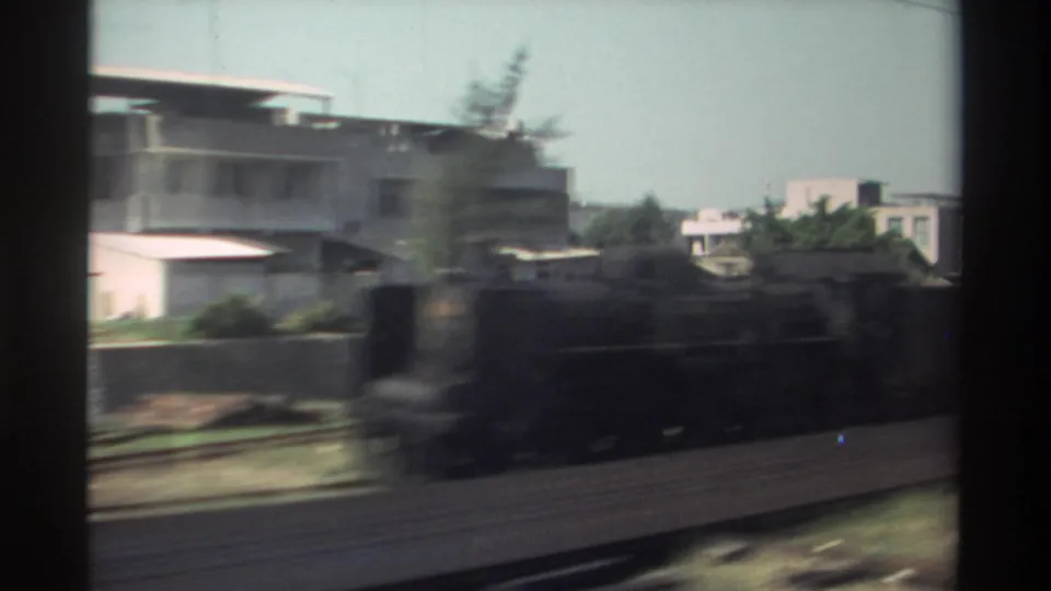 1982 Taiwan train-window shot with parallel tracks, low trackside buildings, trees, and dark motion blur crossing the foreground.