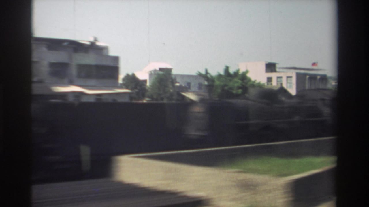 1982 Taiwan train-window view passing low-rise urban buildings, boundary walls, greenery, and tracks under a pale sky.