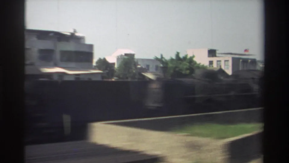 1982 Taiwan train-window view passing low-rise urban buildings, boundary walls, greenery, and tracks under a pale sky.