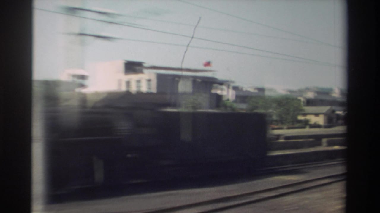 Blurred rail-side view with buildings, tracks, wires, and a dark passing train shape.