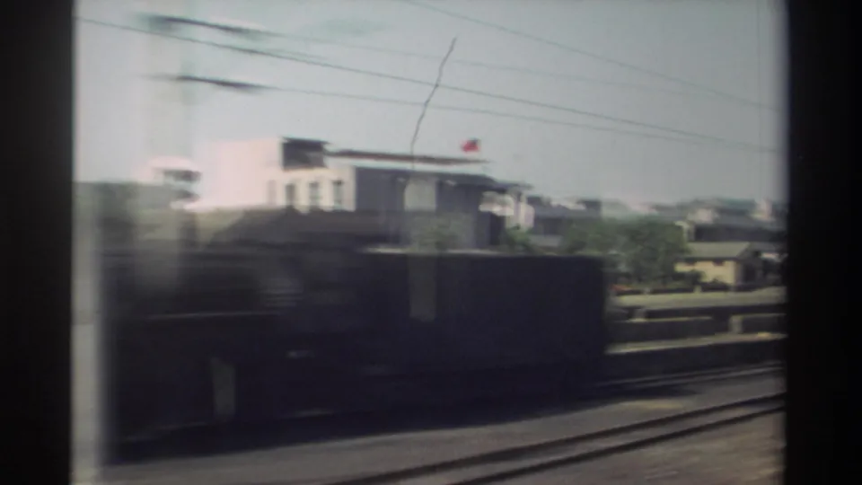 1982 Taiwan footage from a moving train shows rail tracks, low-rise buildings, overhead wires, and a blurred dark rail car or structure.