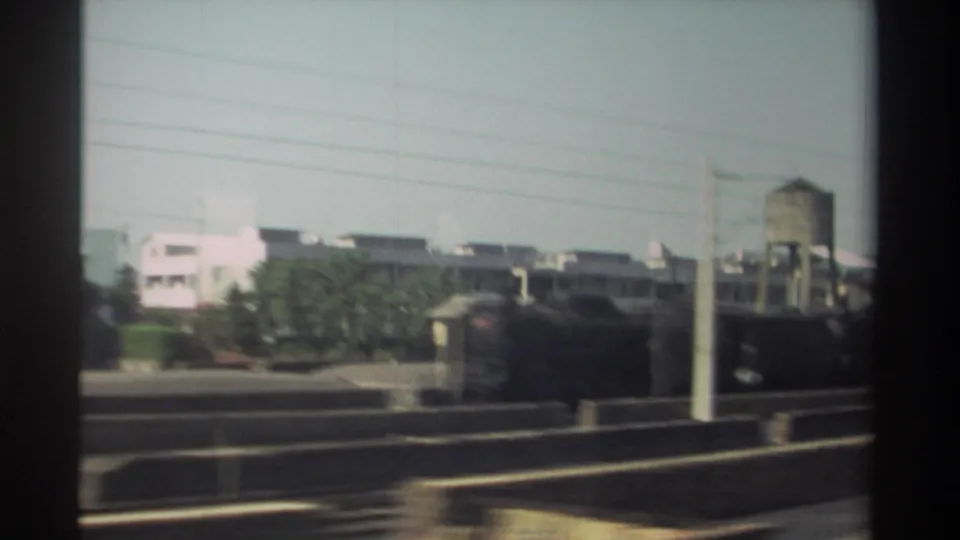 1982 Taiwan train-window view shows low-rise buildings, trees, rail infrastructure, utility wires, and an elevated water tower in the distance.