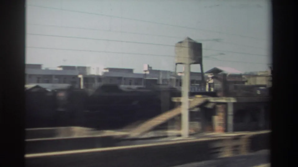 1982 Taiwan urban rail view with an elevated water tower, parallel tracks, low buildings, utility wires, and motion-blurred train cars.