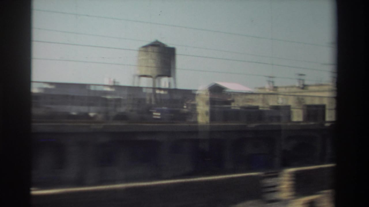 1982 Taiwan train-window view of rail-side industrial buildings, overhead wires, and a prominent water tower.