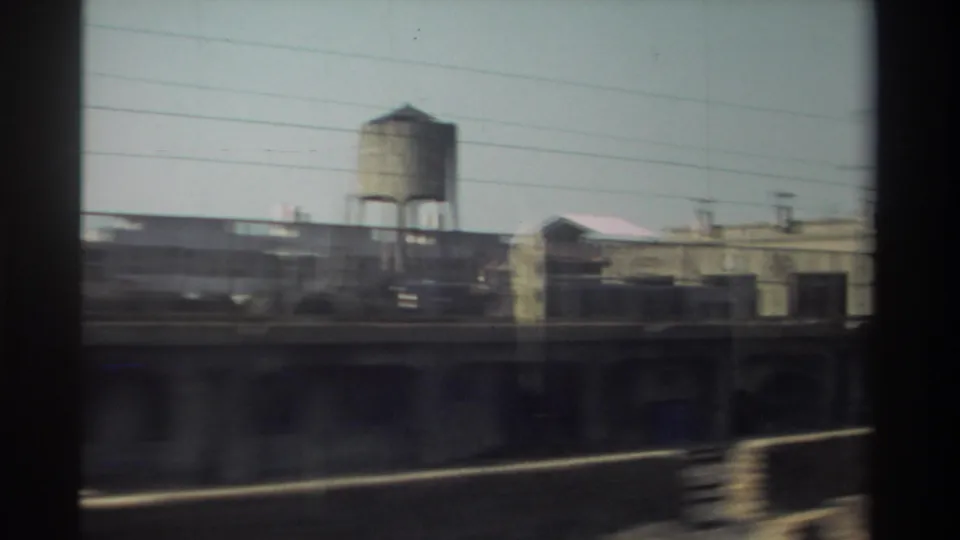 1982 Taiwan train-window view of rail-side industrial buildings, overhead wires, and a prominent water tower.