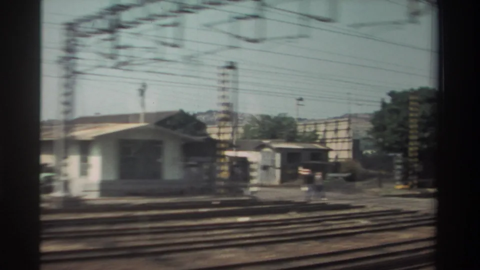 1982 Taiwan passenger-train view with a small rail-side structure, multiple tracks, poles, overhead wires, and distant hillside.