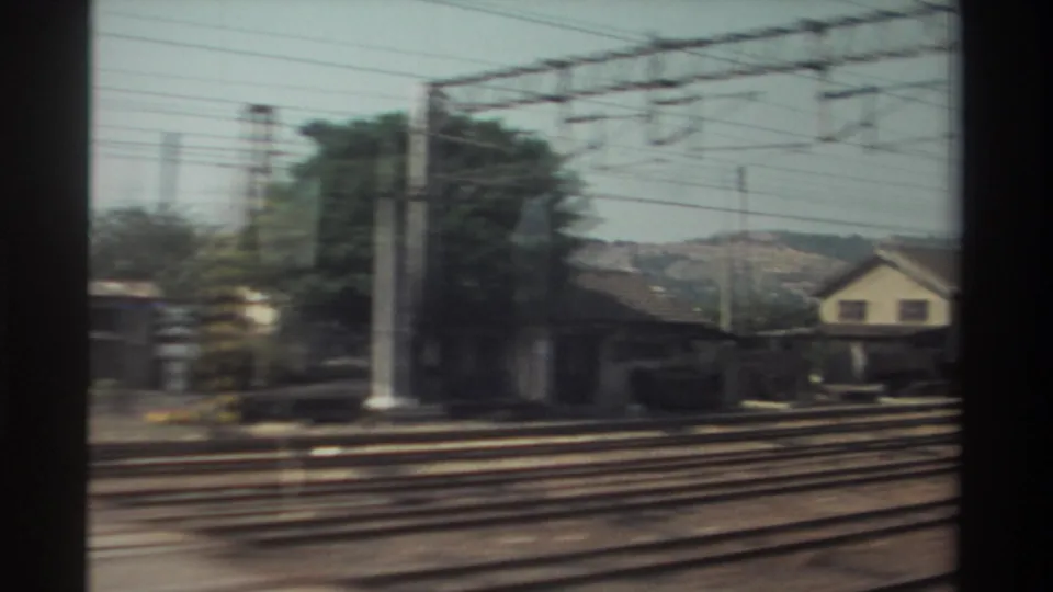 1982 Taiwan train-window frame showing trackside houses, trees, utility poles, overhead lines, and foreground rails in motion blur.