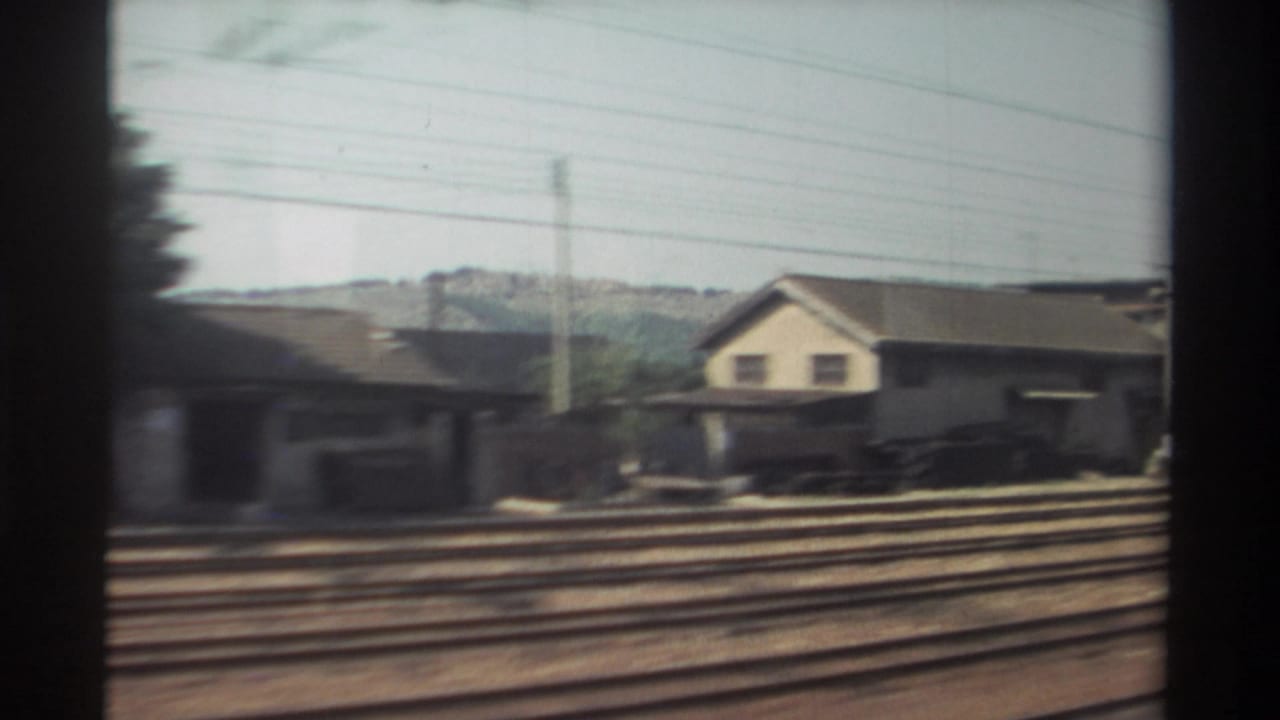 1982 Taiwan home-movie frame showing rural railway tracks, low trackside buildings, utility lines, and distant greenery from a moving train.