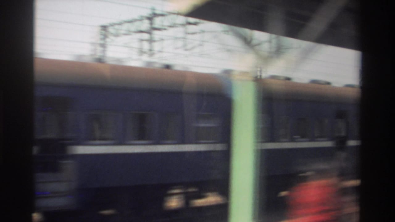 1982 Taiwan 8mm frame of a blue passenger train moving past station columns and overhead rail wires, with heavy motion blur.