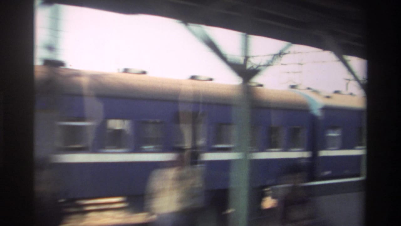 Blue train at a station platform seen through dark framing and overhead wires.