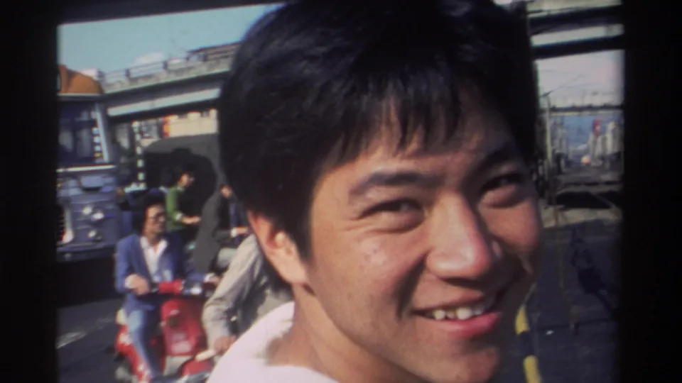 1982 Taiwan home-movie frame of a smiling young man in close-up near a busy station area with commuters, vehicles, and a bridge behind him.