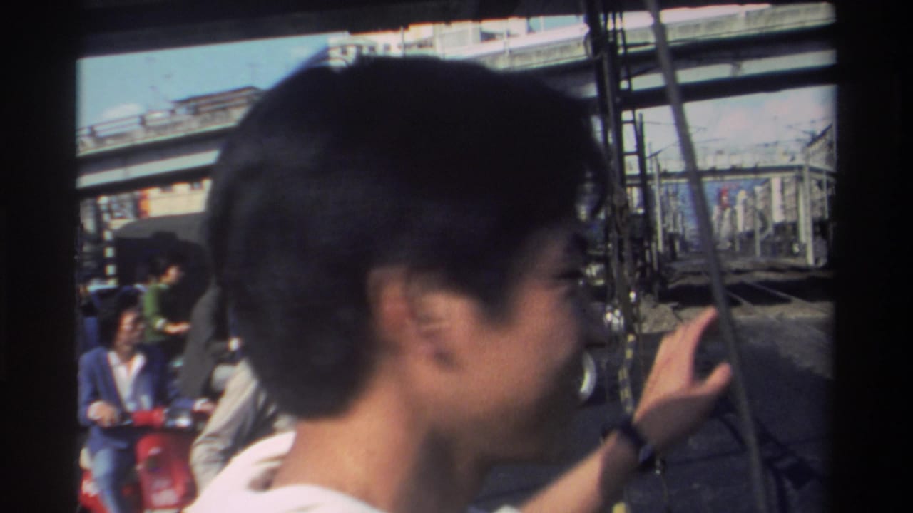 Young man in profile raises a hand on a busy street with scooters and elevated structures behind him.