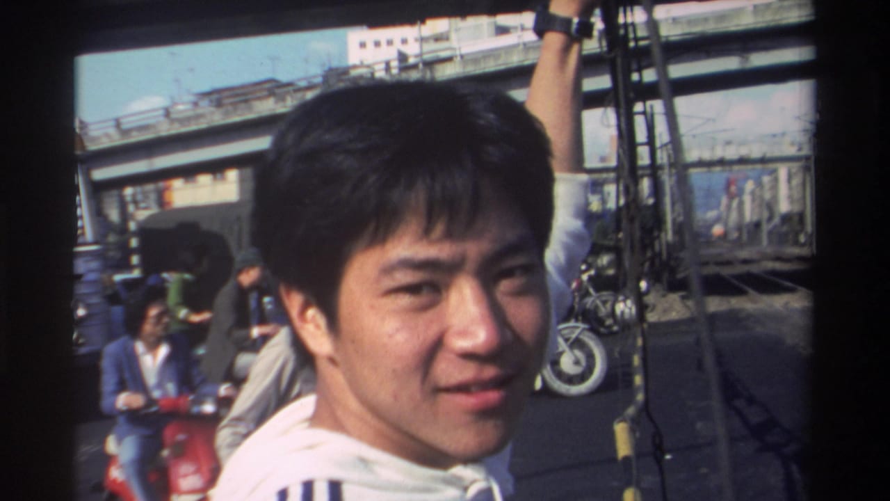Young man faces the camera and smiles on a street with scooters, pedestrians, and elevated tracks behind him.