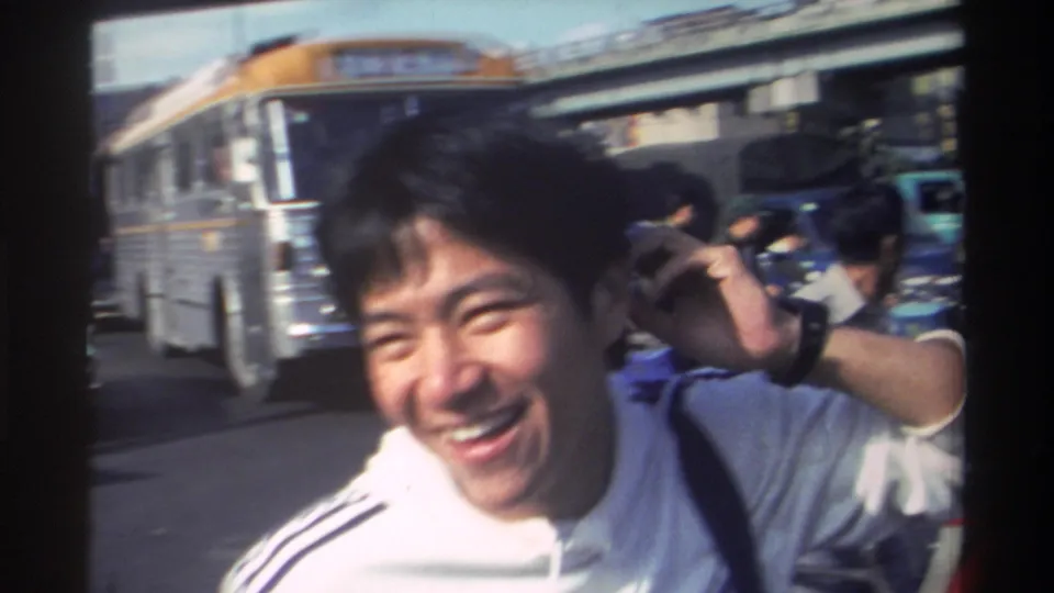 The young man laughs and lifts his hand toward the camera in 1982 Taiwan, with a bus, scooters, pedestrians, and urban street structures nearby.