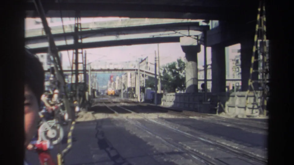 1982 Taiwan home-movie frame with a face at the left edge, rail lines, concrete supports, and a yellow train in the distance.
