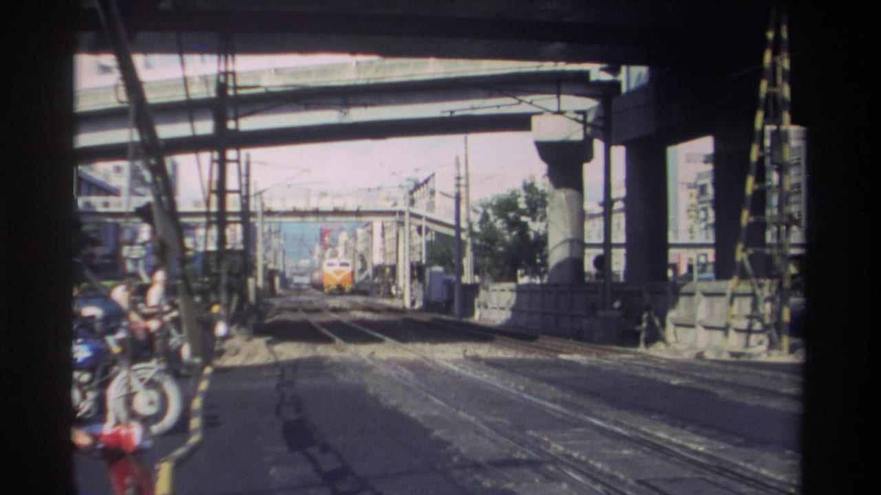 1982 Taiwan home-movie frame showing an orange and white train approaching under concrete bridge structures beside overhead rail wires.
