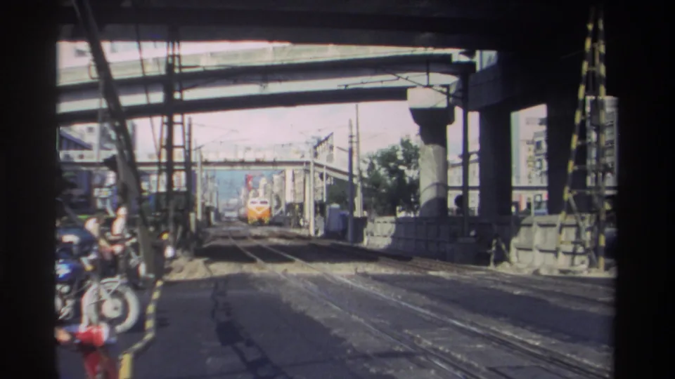 1982 Taiwan home-movie frame showing an orange and white train approaching under concrete bridge structures beside overhead rail wires.