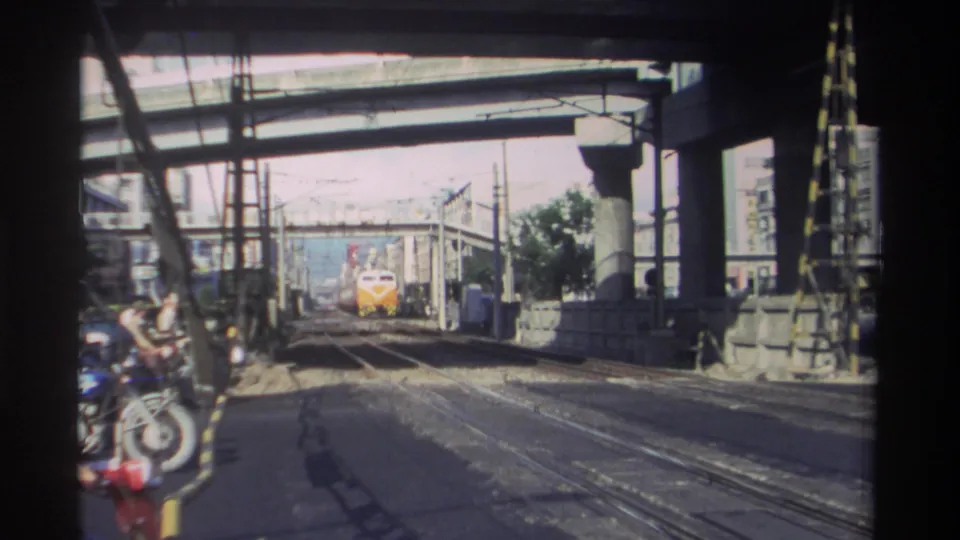 Approaching train in Taiwan, 1982, framed by concrete overpass spans, overhead wires, rail tracks, and roadside industrial details.