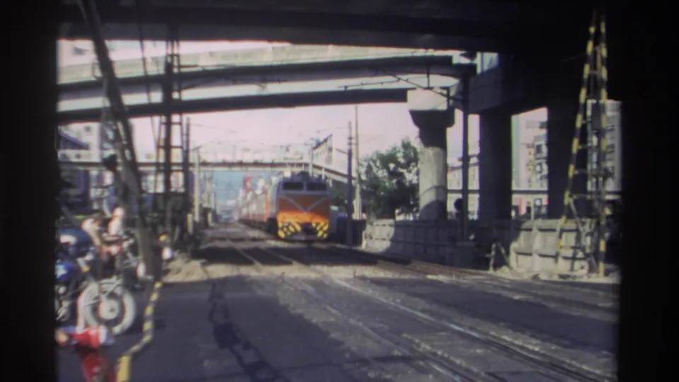 Orange-fronted train nears the camera under a concrete overpass, with rail lines, support columns, and industrial surroundings visible in Taiwan, 1982.