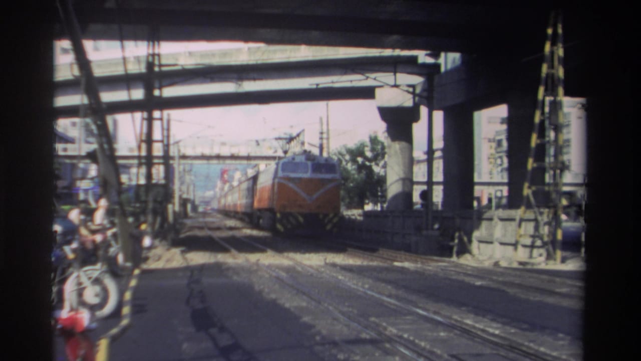 1982 Taiwan home-movie frame showing an orange and white passenger train approaching beneath concrete overpasses and overhead wires.