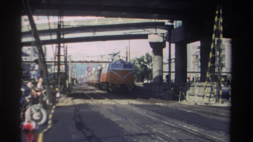 1982 Taiwan home-movie frame showing an orange and white passenger train approaching beneath concrete overpasses and overhead wires.