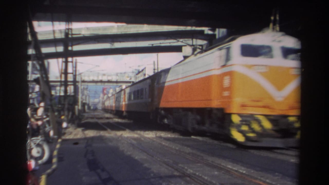 Train nose passes close to camera beneath overpasses, with rails and motorcycles visible.