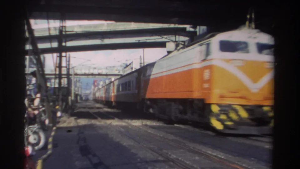 Orange and white passenger train speeds past the camera in Taiwan, framed by a dark foreground and urban rail infrastructure.