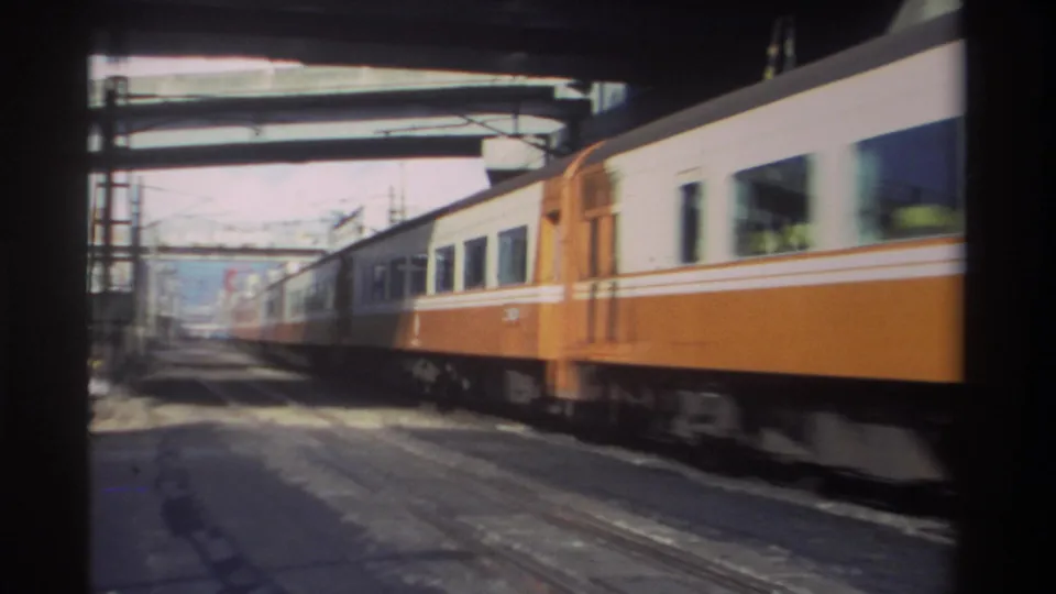 Orange and white passenger cars recede through an urban rail underpass in Taiwan, with tracks, wires, and shadowed framing visible.
