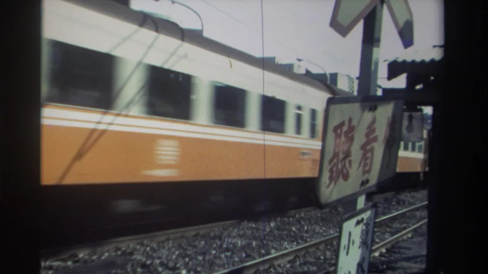 1982 Taiwan home-movie still of a passenger train car passing behind warning signage, with rail ballast and dark frame edges visible.