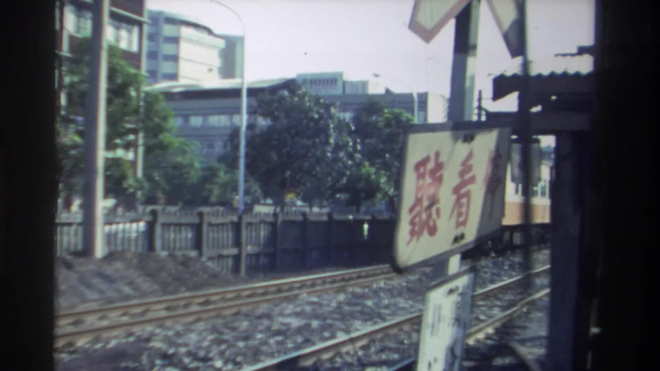 After the train passes, tracks, trees, urban buildings, and a Chinese-character crossing sign remain in view in Taiwan.