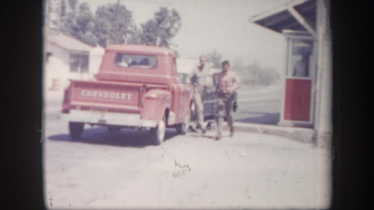 Red Chevrolet pickup parked near a small roadside building with adults standing beside it.