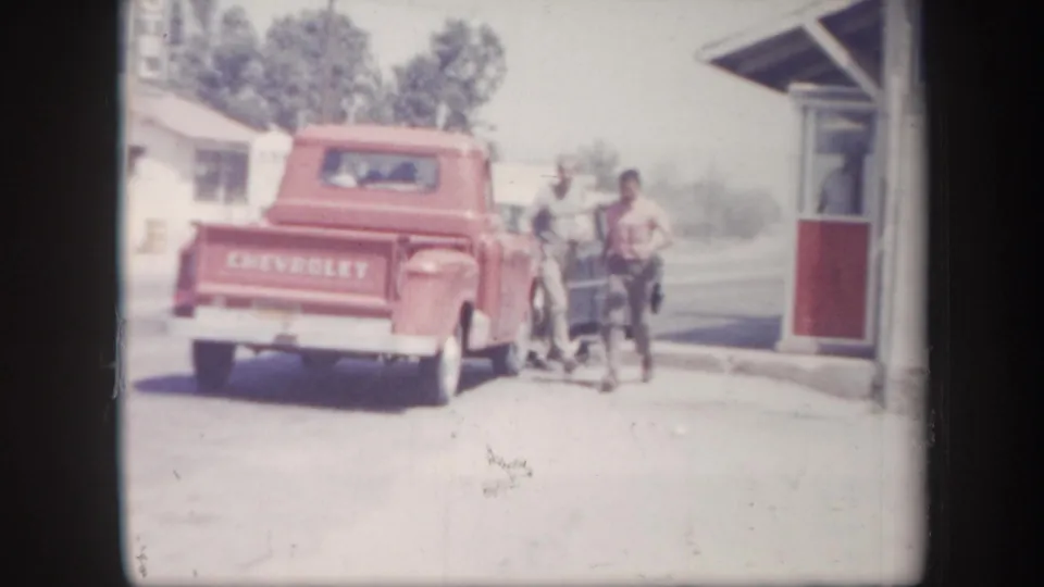 1957 Arizona home-movie frame showing a red Chevrolet pickup, adults nearby, and a small roadside building in bright daylight.