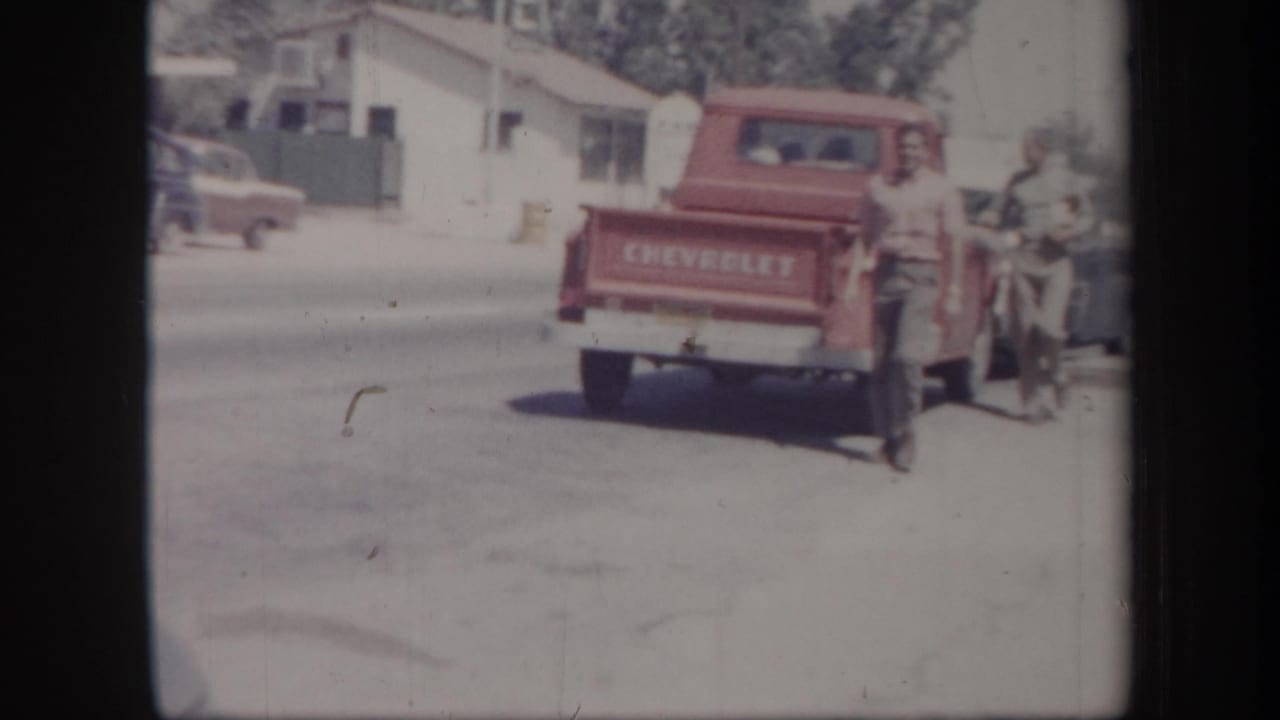 1957 Arizona frame with a red Chevrolet pickup on a quiet street, people nearby, and mid-century cars and buildings in the background.