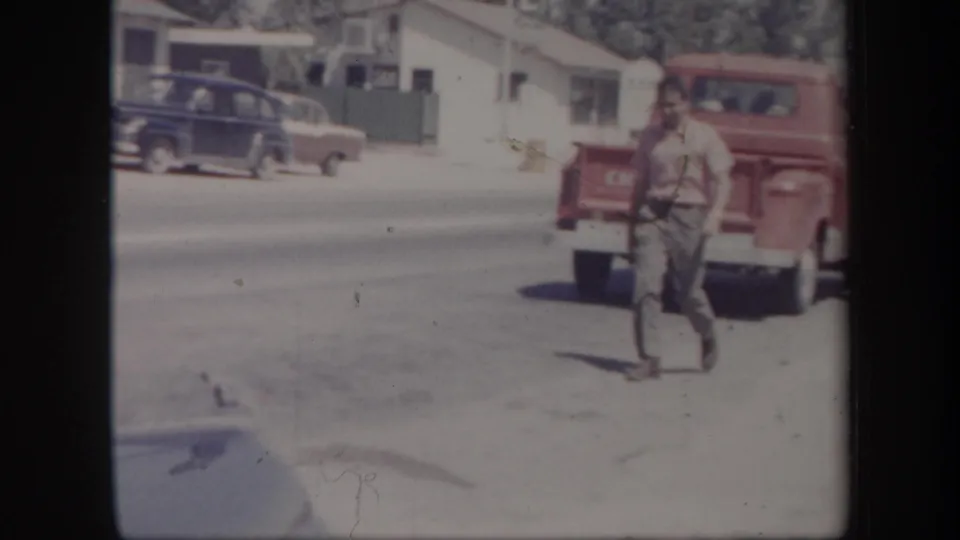 1957 Arizona home-movie frame showing a man in a red shirt moving past a parked red Chevrolet pickup on a bright street.