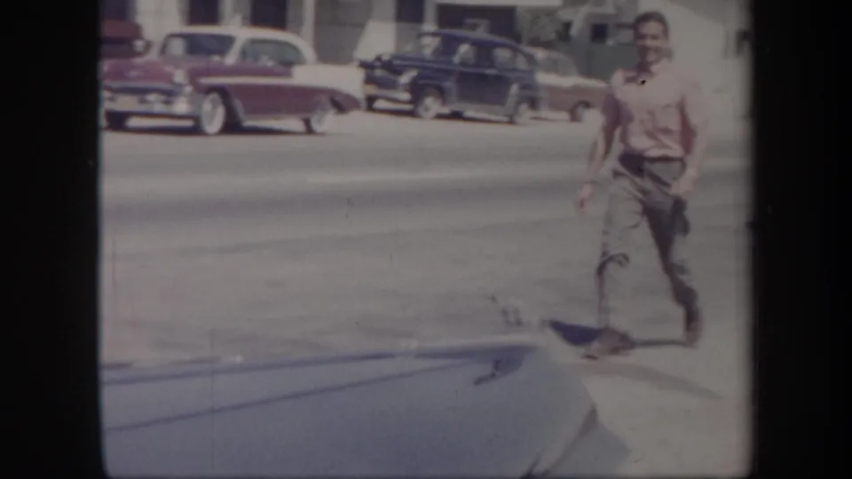 1957 Arizona frame showing a man in a red shirt near the camera, with mid-century cars parked across the sunlit street.