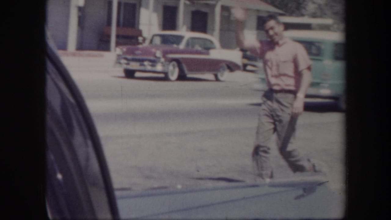 Man in a pink shirt waves near a street with classic cars passing behind him.