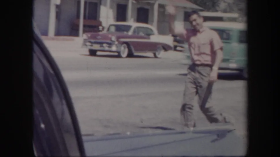 1957 Arizona home-movie frame showing a man waving on a sunlit street as mid-century cars pass in the background.