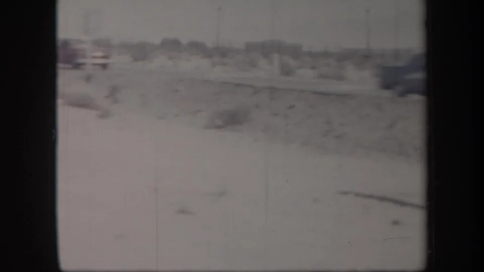 1957 home-movie frame showing a washed, handheld view of desert roadside terrain with sparse brush and distant poles.