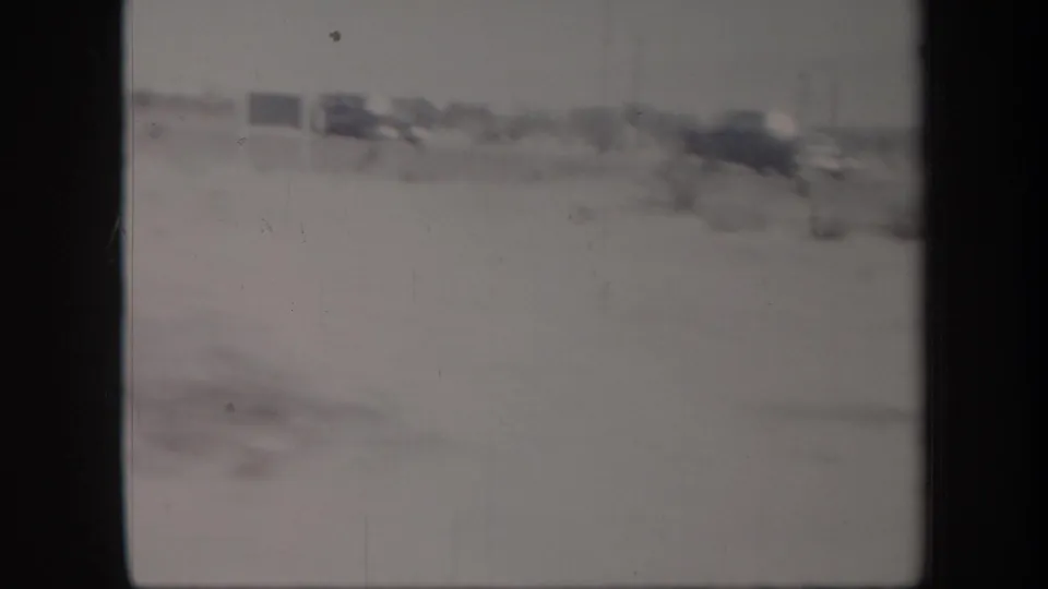 Handheld 8mm frame of a desert roadside with distant signs, scrub, and dark vehicles or roadside objects at right.