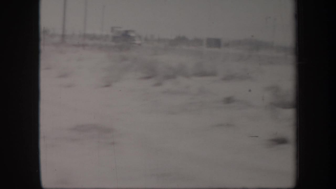 Soft, blurred 1957 Arizona desert view with sparse scrub and distant roadside forms on faded 8mm film.