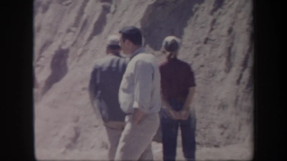 Adults in mid-century casual clothing stand near a rocky Arizona roadside slope during a travel stop.