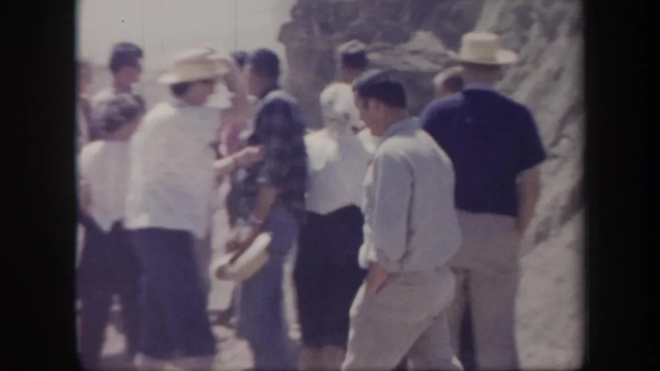 A group of adults in 1950s summer clothing gathers near a rocky Arizona roadside during a road-trip break.