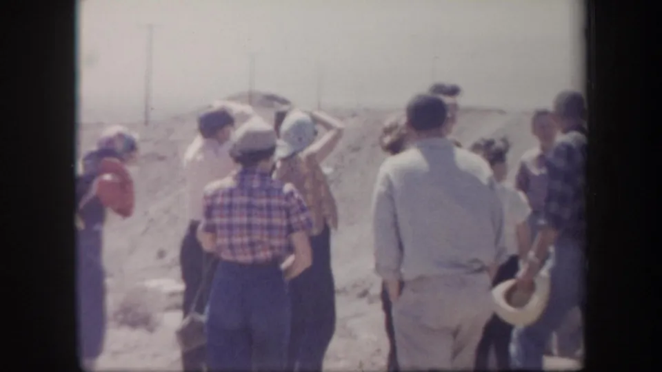 Adult travelers in hats and casual 1950s clothing stand along a dusty Arizona roadside near rocky terrain.