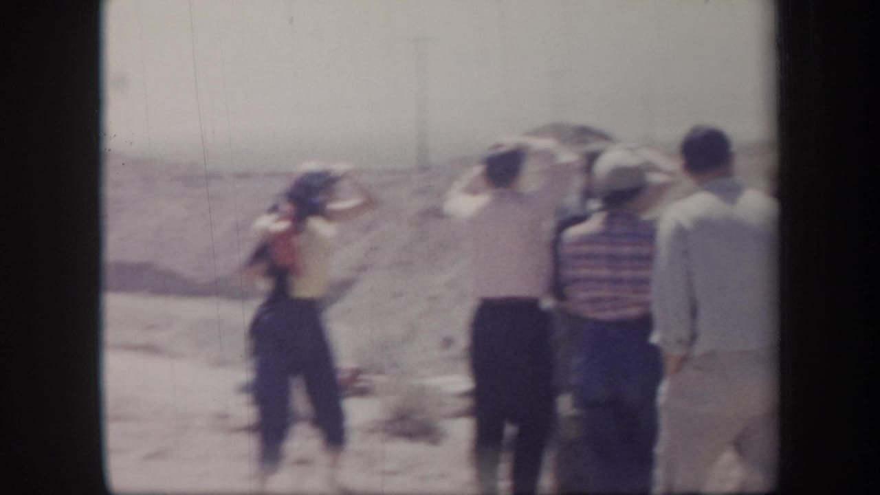 1957 Arizona 8mm frame of adults gathered on a dusty desert slope, several facing away from the camera in bright sunlight.
