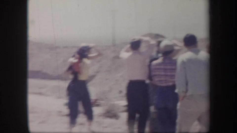 1957 Arizona 8mm frame of adults gathered on a dusty desert slope, several facing away from the camera in bright sunlight.
