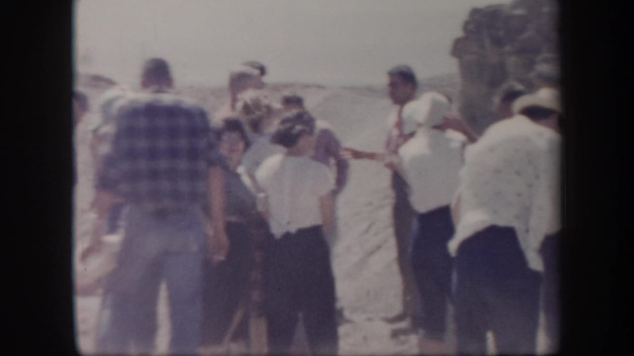 A group of adults stands in a dusty open area near a rocky edge.