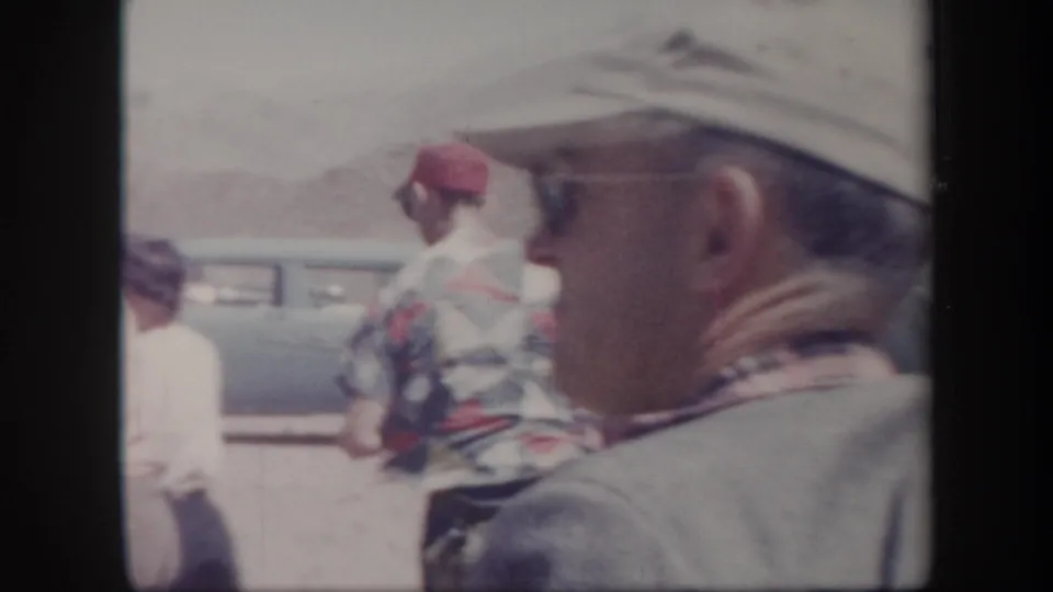 Soft 8mm frame with a close adult profile in the foreground, people nearby, and a parked car in the Arizona sun.